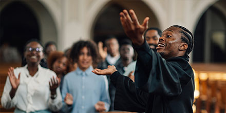 A person in a robe sings passionately with an outstretched arm, while a group of people stand behind, clapping and smiling in a church setting.
